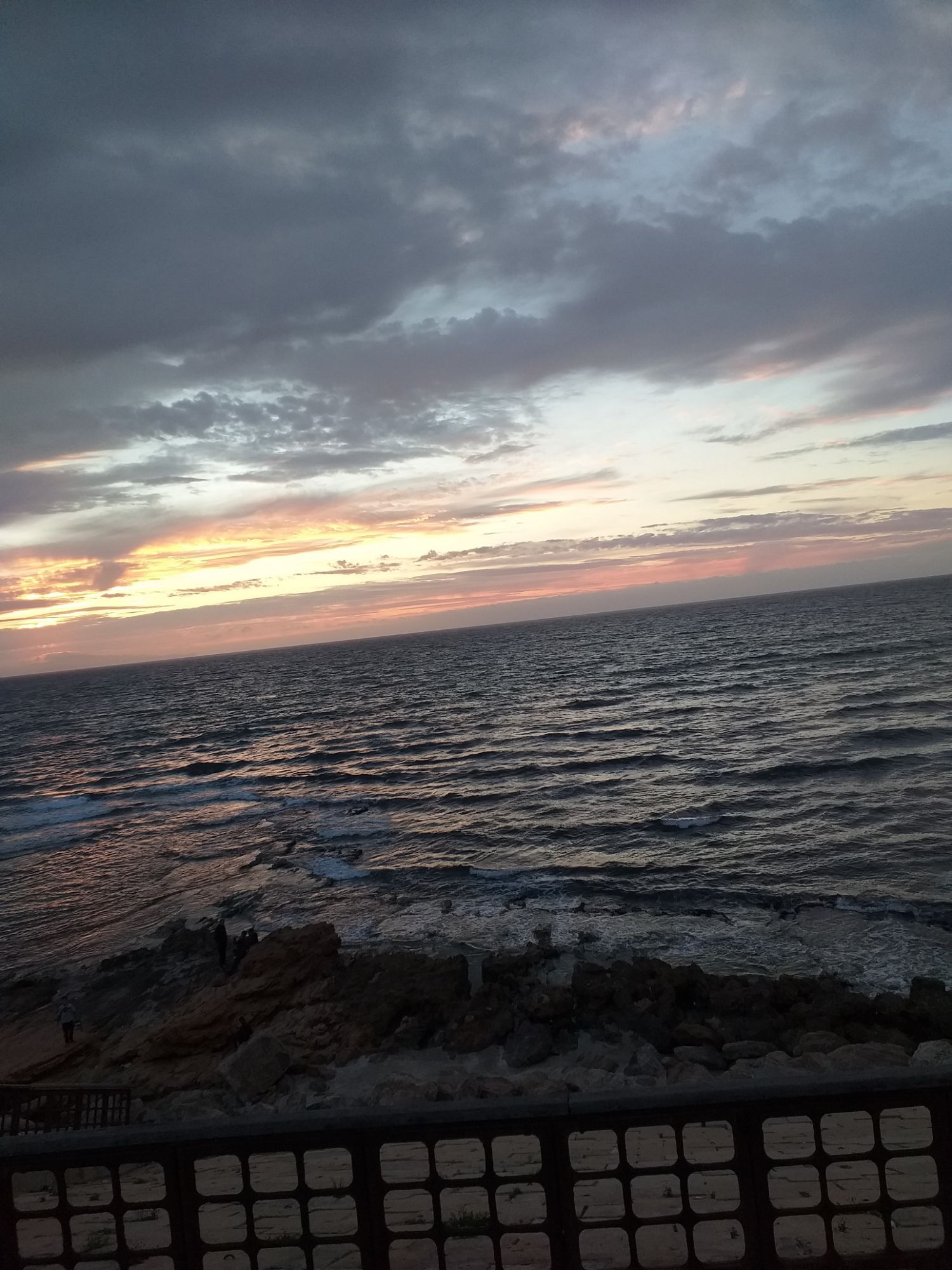 Coucher de soleil sous un ciel nuageux, au-dessus d’une mer agitée et de rochers, vu depuis une balustrade