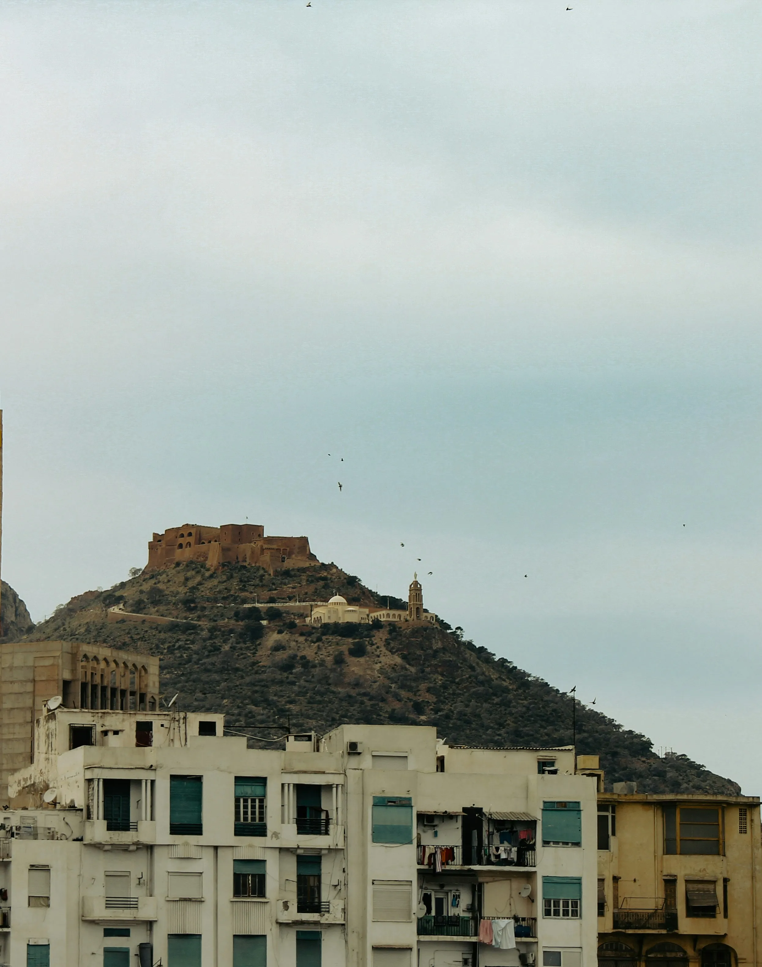 Au-dessus d’immeubles clairs, une colline couverte de broussailles ; au sommet, une forteresse de pierre ocre, et plus bas sur le versant, une coupole blanche et une tour
