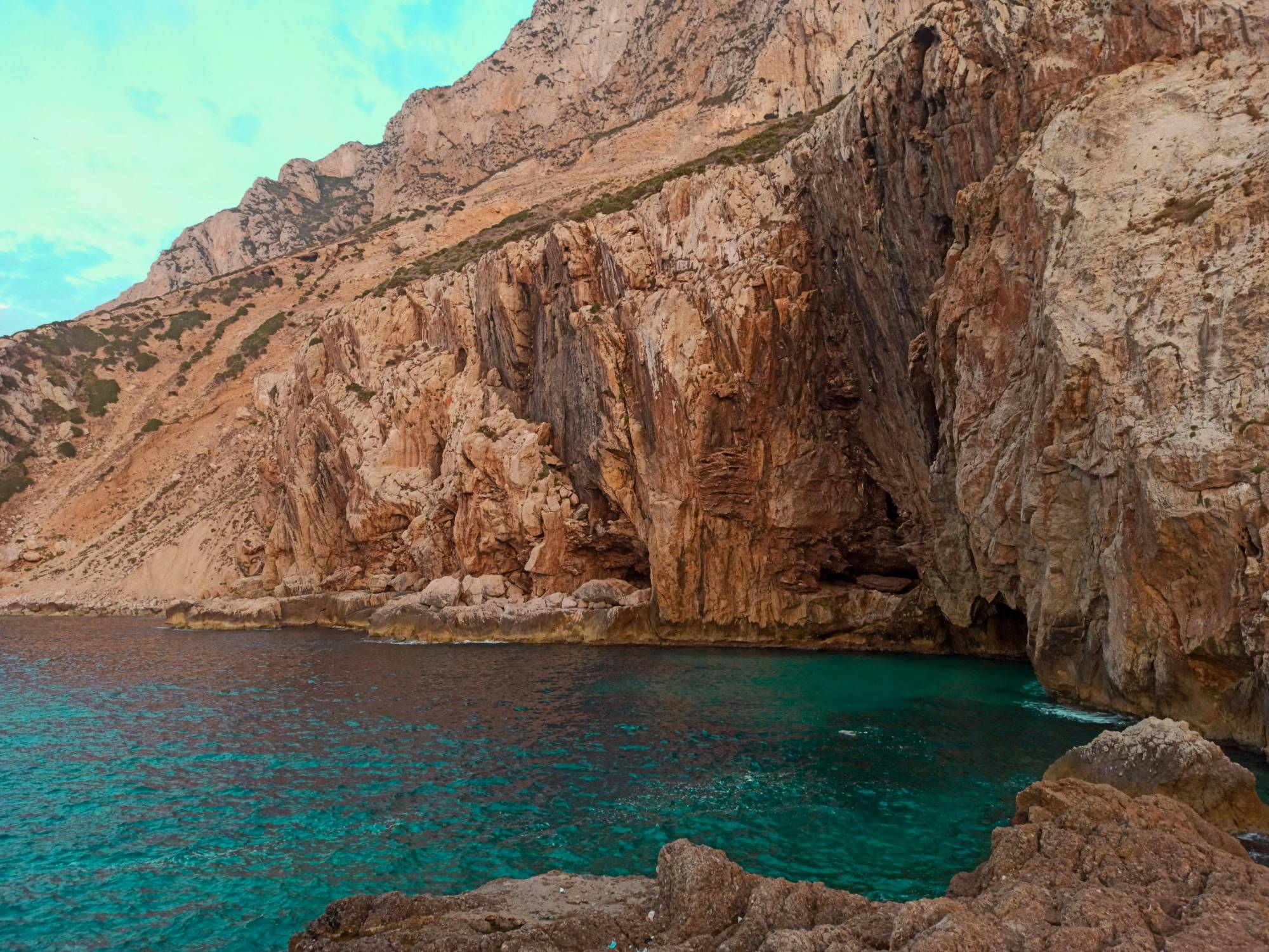 Hautes falaises de roche ocre et rouille plongeant dans une crique d’eau turquoise, avec l’entrée sombre d’une grotte au pied de la paroi