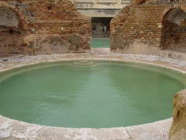 Un bassin circulaire rempli d’une eau vert pâle, entouré d’une margelle de pierre, entre des murs antiques de brique et de pierre percés d’arcs