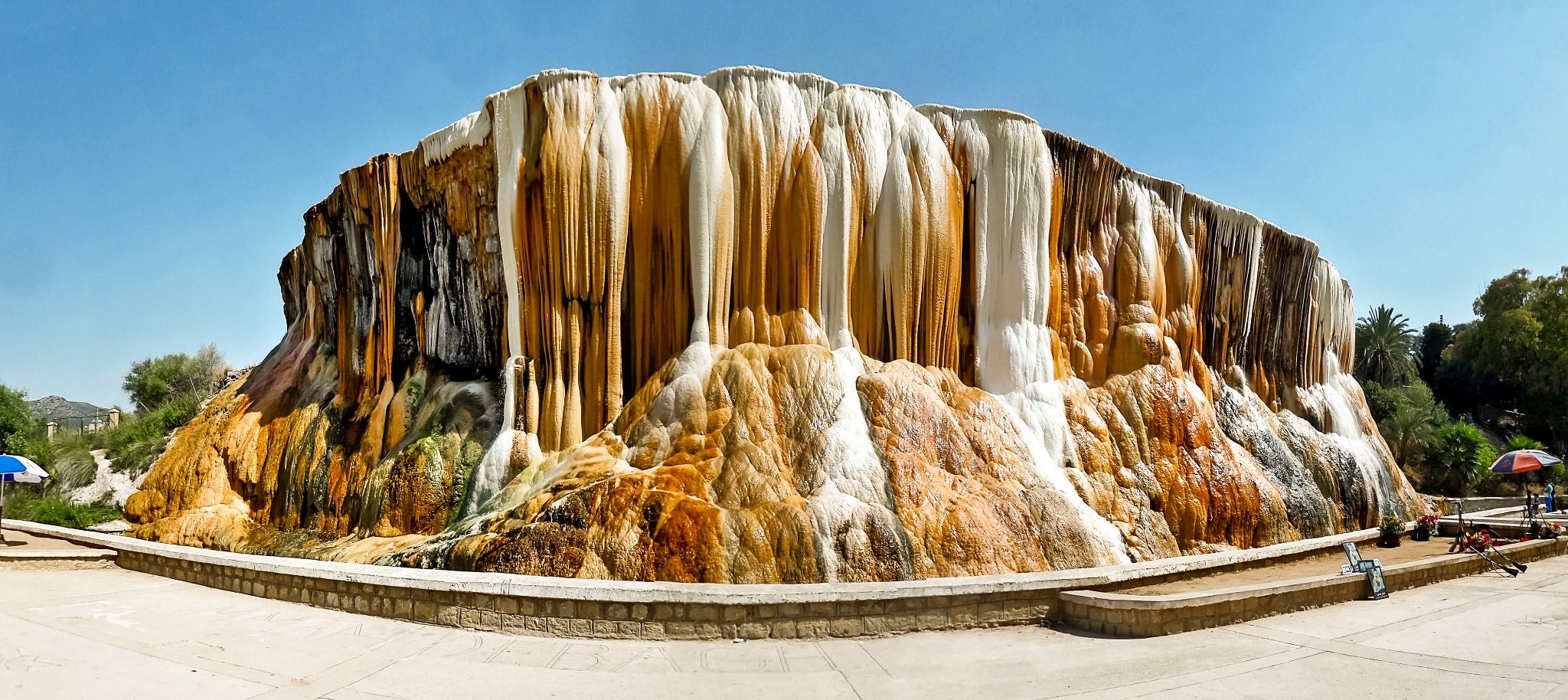 La cascade de Hammam Debagh en entier : un large massif de calcaire strié de blanc, d’ocre et de brun, posé au bord d’une esplanade pavée, sous un ciel bleu