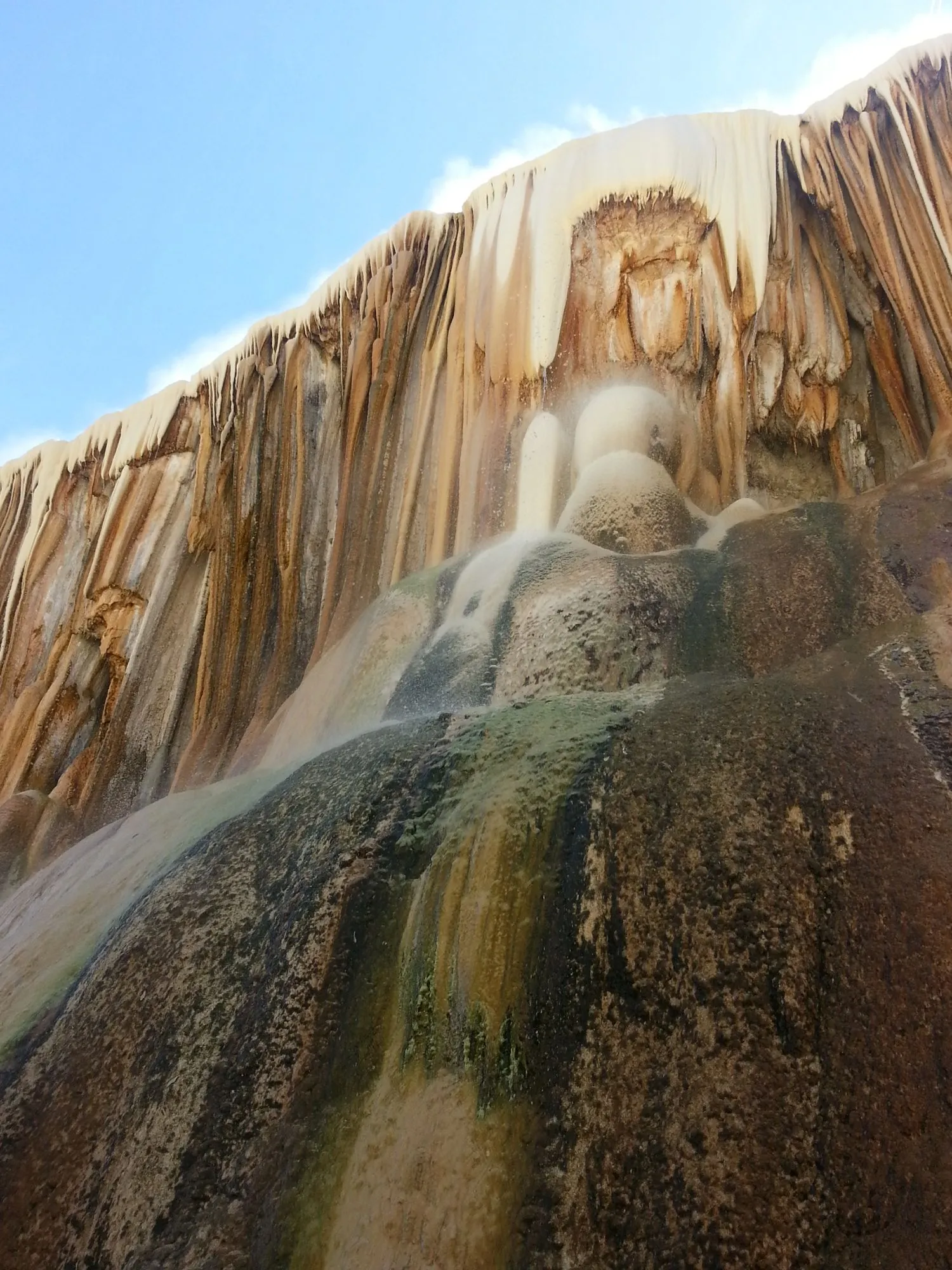 La cascade vue d’en bas : un rideau de calcaire strié de blanc, d’ocre et de brun, sur lequel ruisselle de l’eau, sous un ciel bleu