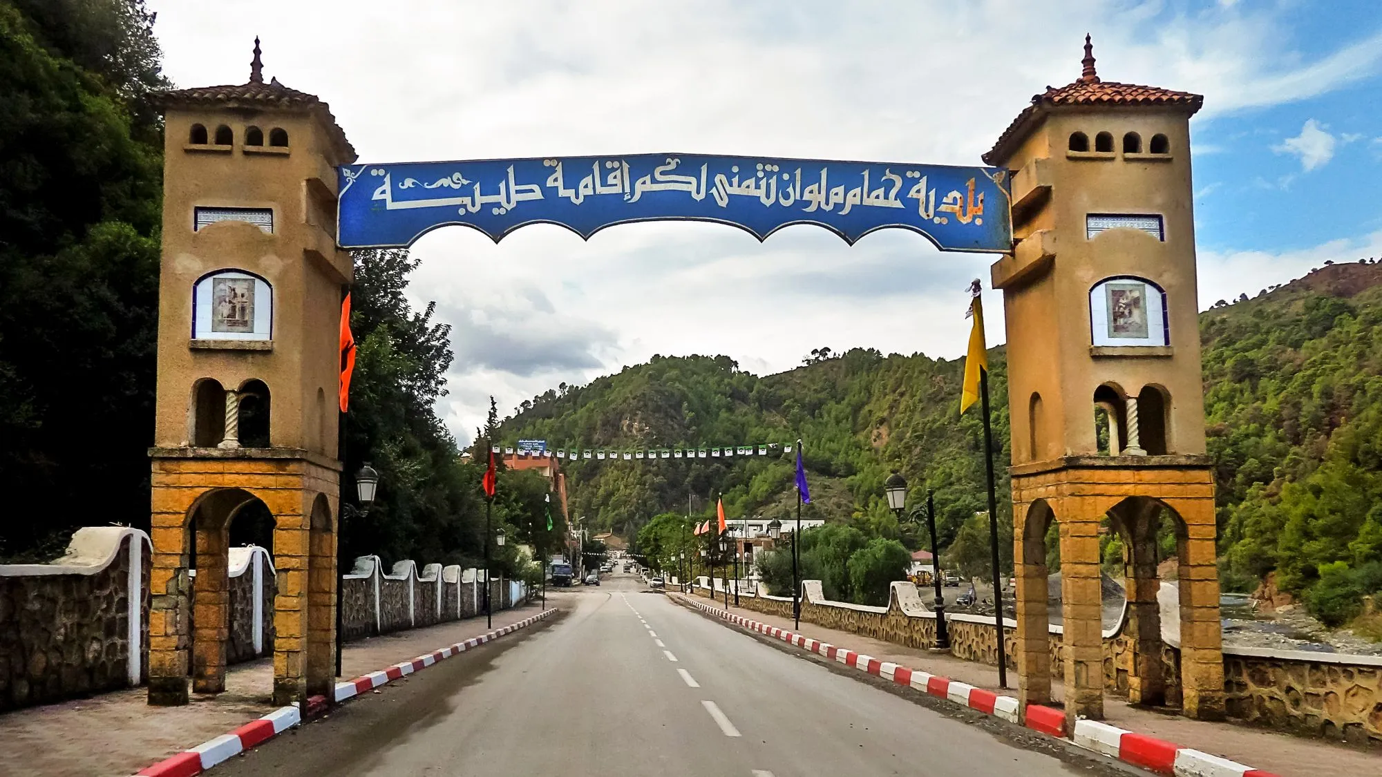 Une route passe sous un portique bleu inscrit en arabe, entre deux tours ocre à arcades, et s’enfonce dans une vallée aux collines boisées