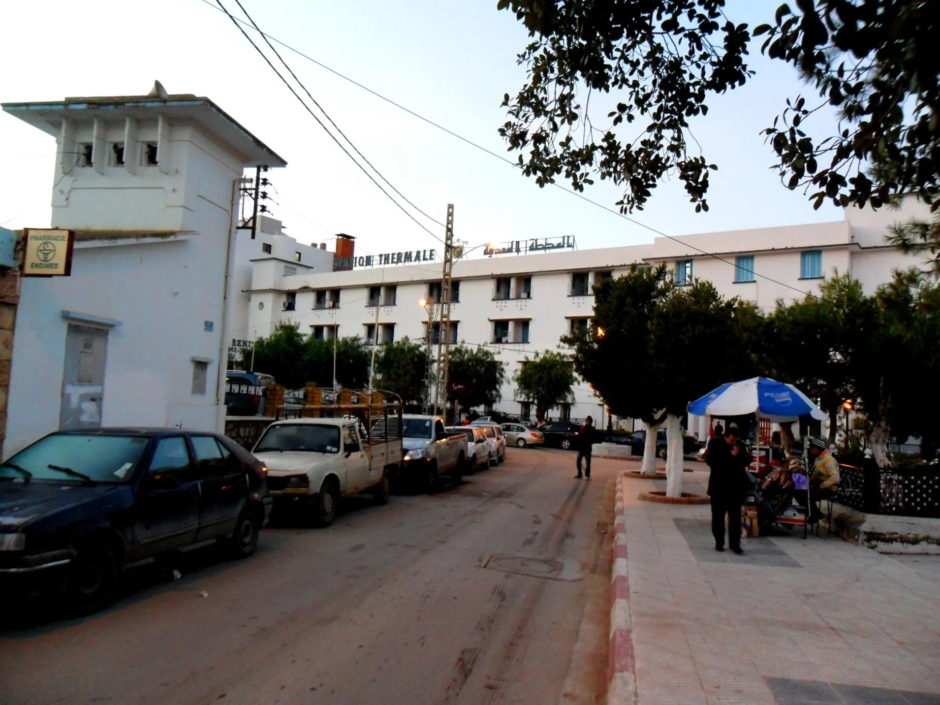 Une rue au crépuscule, bordée de voitures garées, devant un long bâtiment blanc de plusieurs étages surmonté de l’inscription « Station thermale » en français et en arabe
