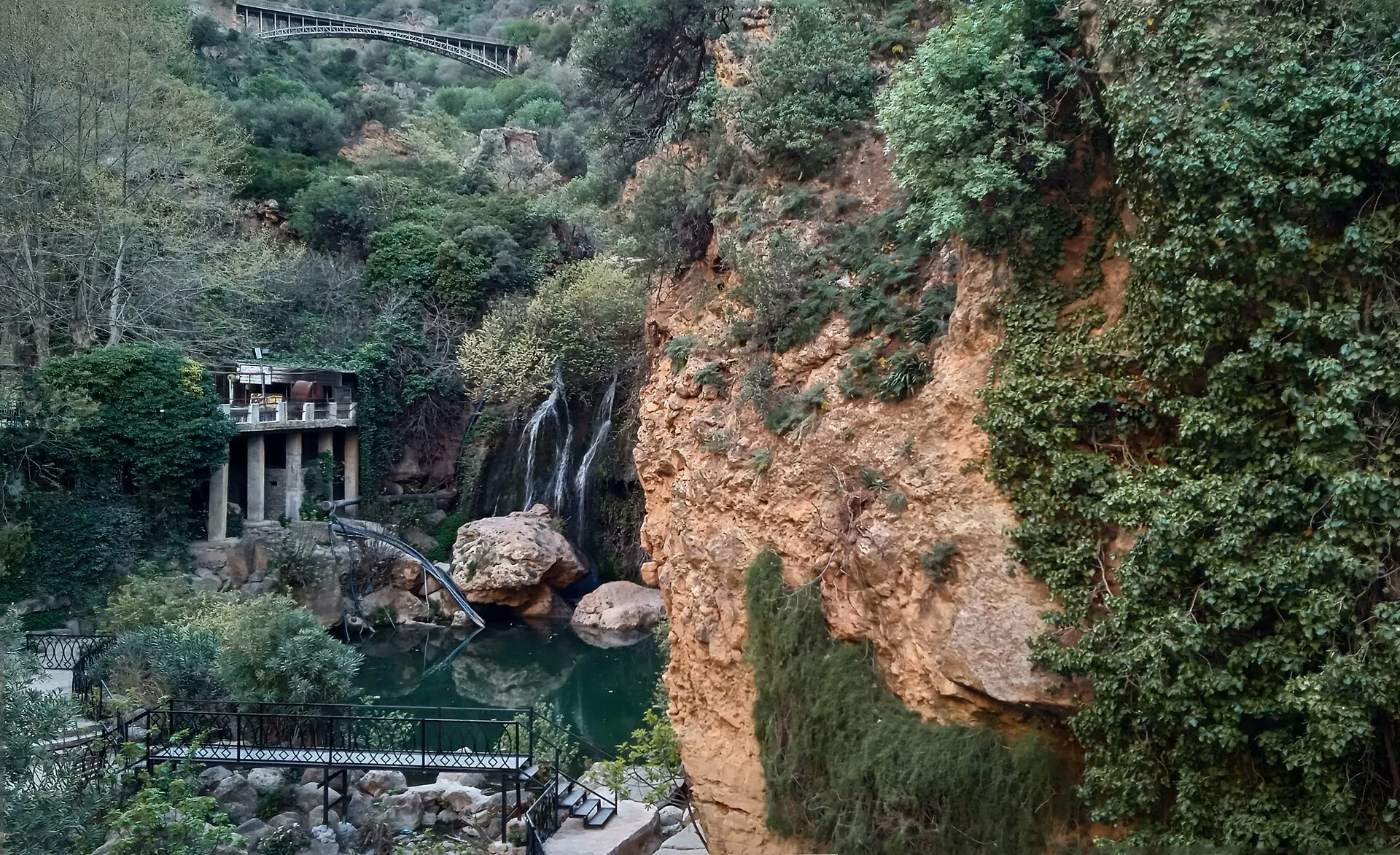 Une cascade tombe entre des rochers dans un ravin couvert de verdure, au pied d’une falaise ocre ; plus haut, un pont franchit la gorge