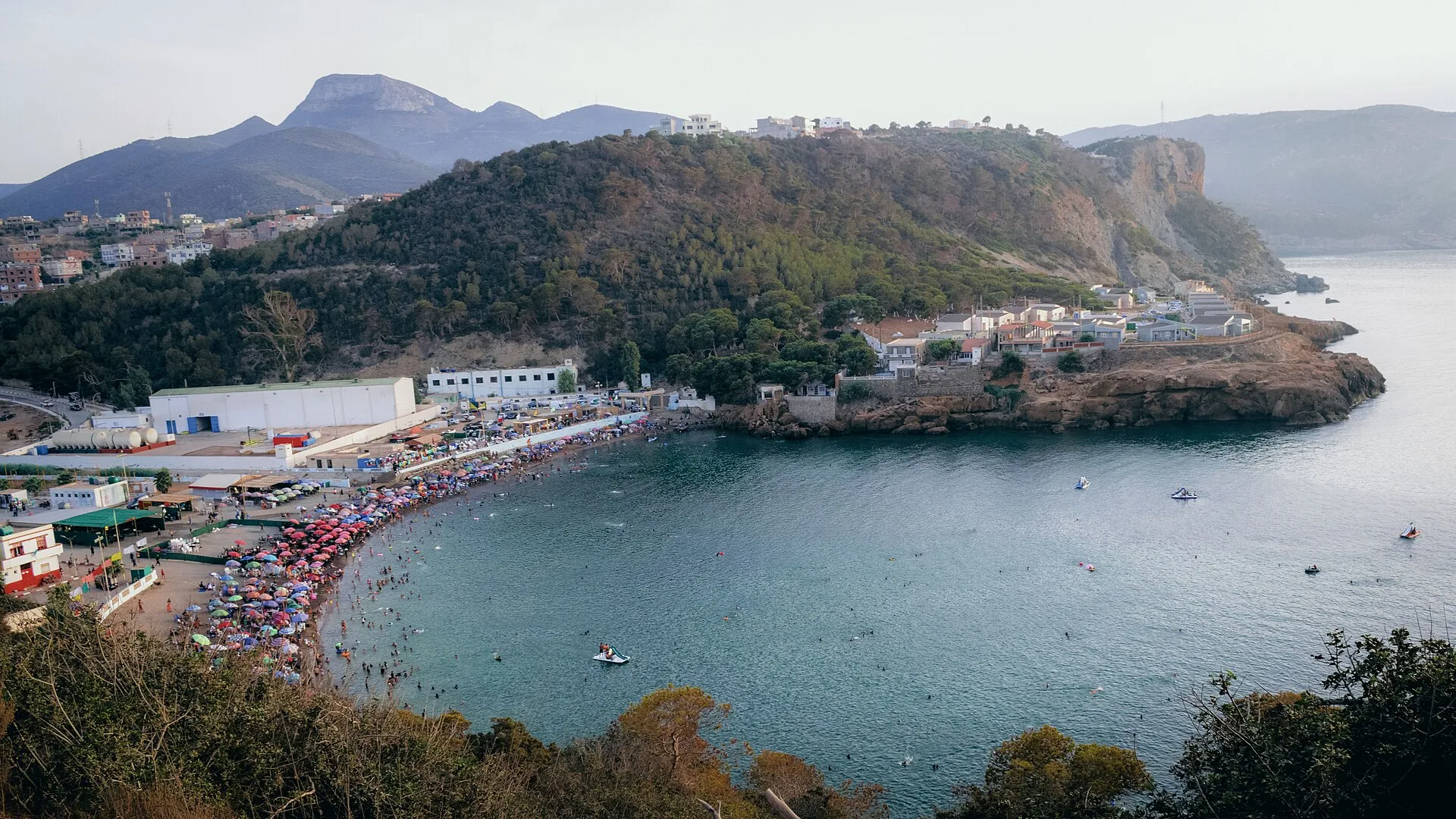 Une baie aux eaux calmes, bordée d’une plage couverte de parasols, au pied d’une colline boisée et de montagnes ; quelques bateaux sur l’eau