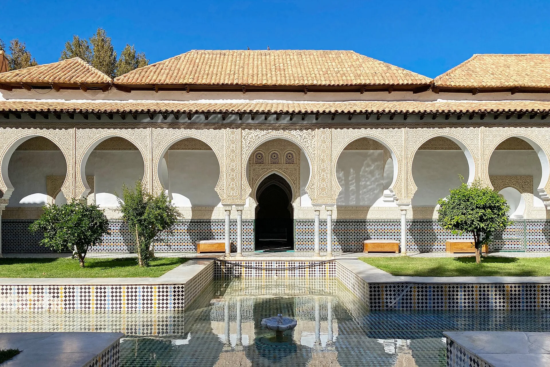 Cour du palais du Mechouar : un long bassin bordé de zelliges, de petits arbres et une galerie d’arcs festonnés en plâtre blanc sous des toits de tuiles