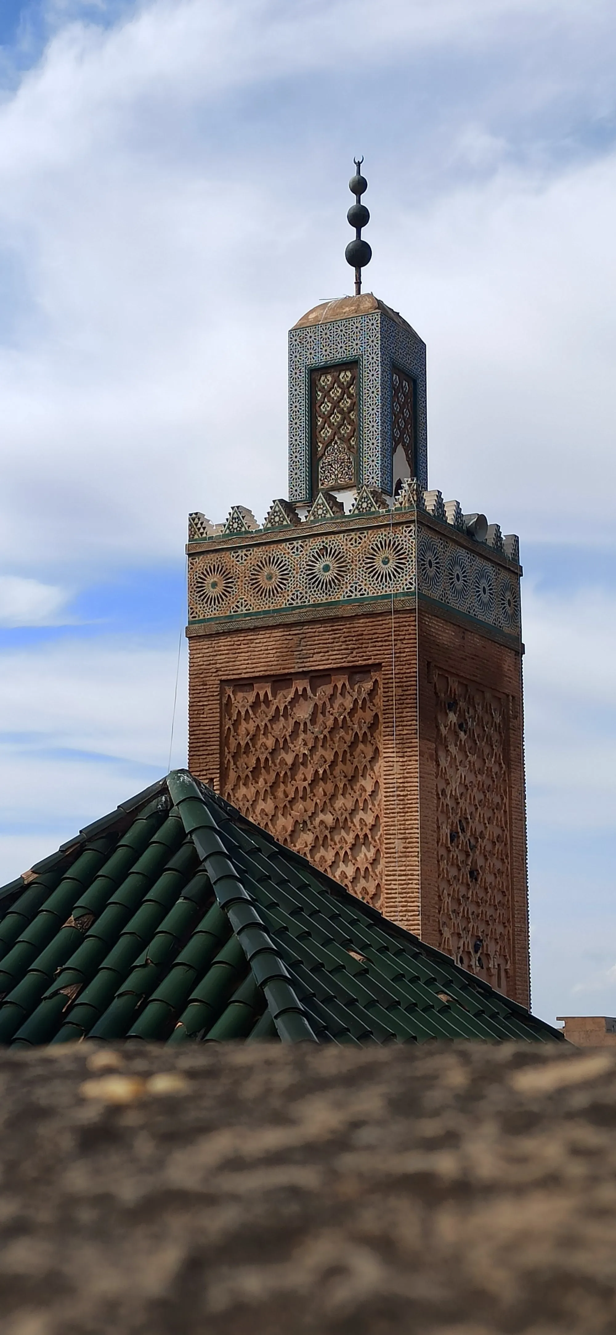 Un minaret carré en brique, orné de panneaux sculptés en losanges et d’une frise de céramique à rosaces, coiffé d’un lanternon carrelé ; au premier plan, un toit de tuiles vertes