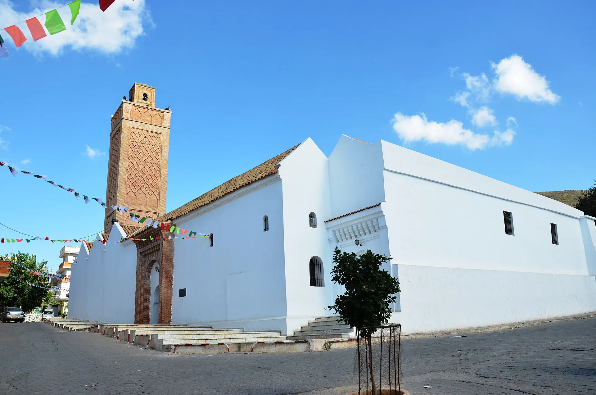 La Grande Mosquée de Nedroma : de longs murs blanchis sous des toits de tuiles et un minaret de brique à décor en losanges, sous un ciel bleu traversé de guirlandes de fanions