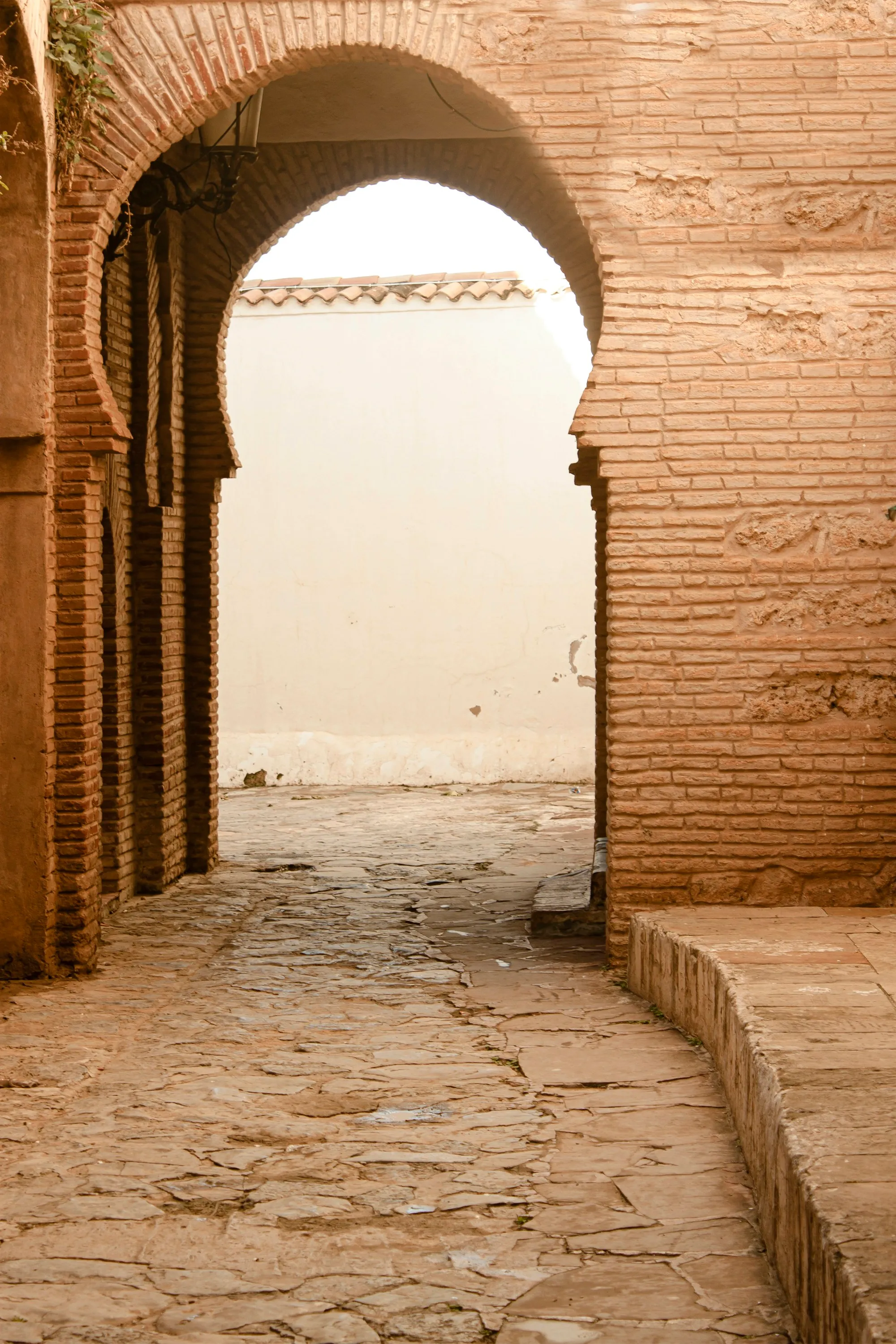 Ruelle dallée de pierres irrégulières qui passe sous un arc outrepassé en brique ocre ; au-delà, un mur blanc coiffé de tuiles, et une lanterne accrochée à gauche