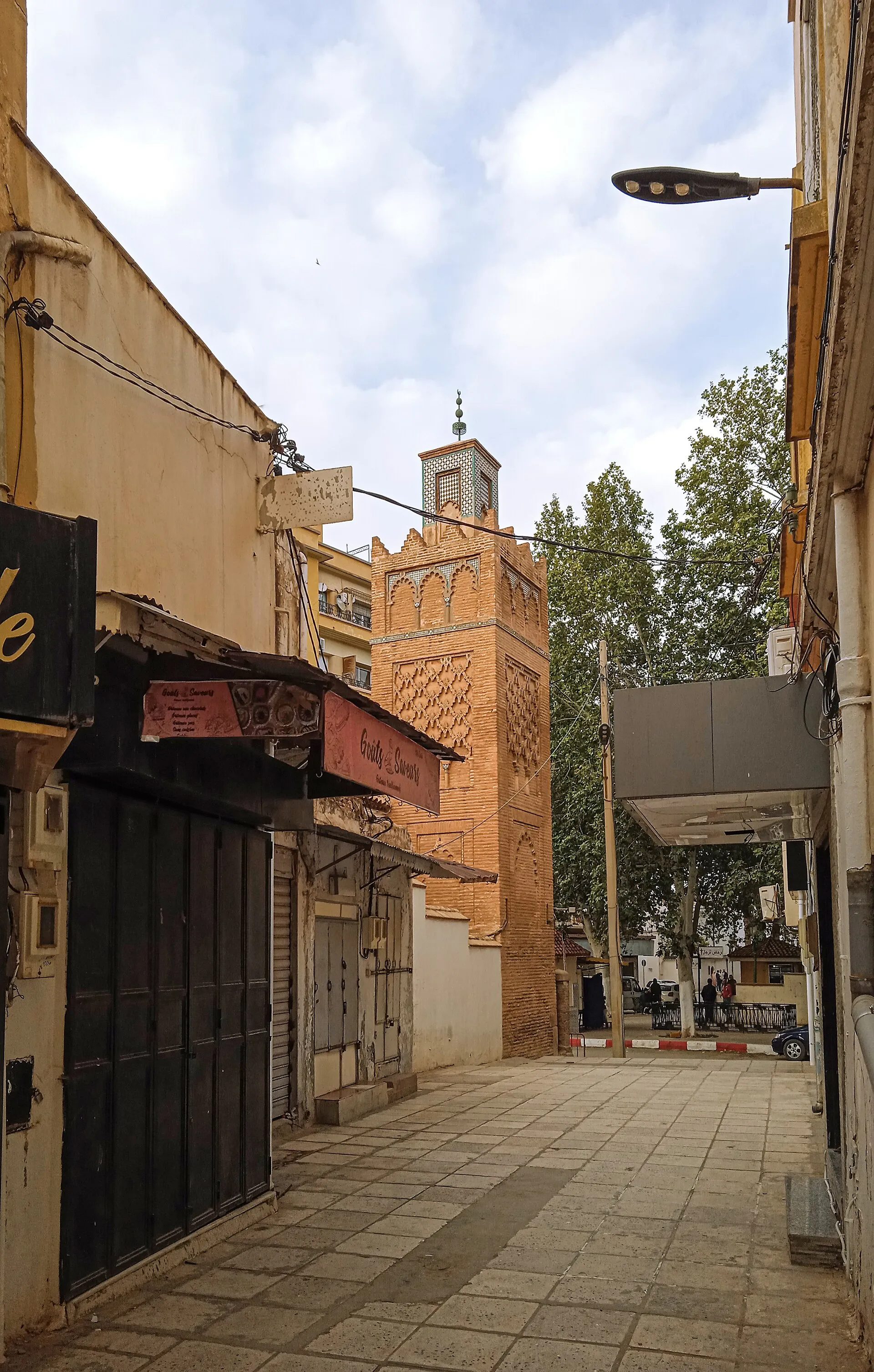 Une rue piétonne étroite, bordée de boutiques aux rideaux baissés ; au bout, un minaret de brique ocre à décor en losanges se détache sur le ciel