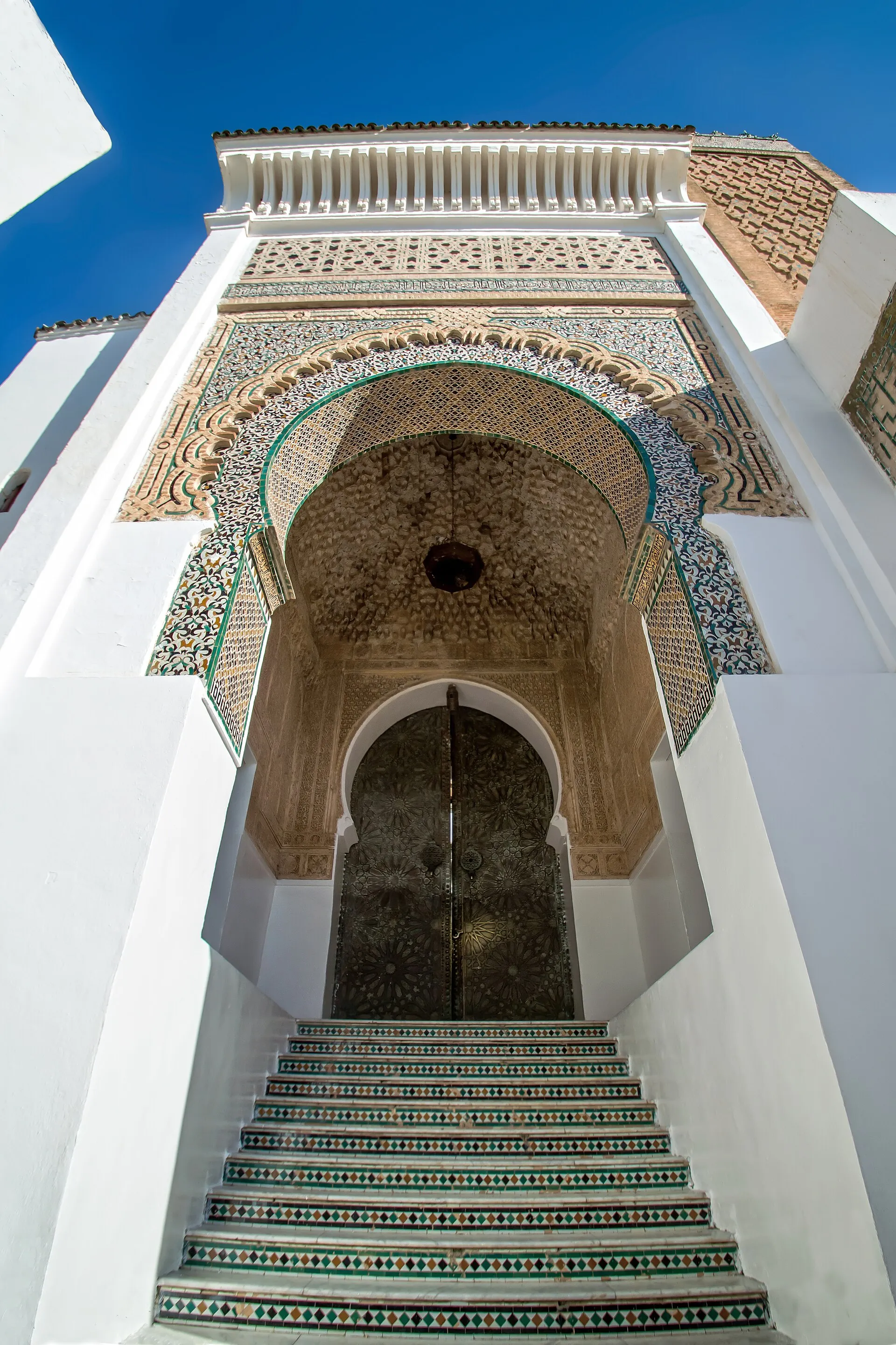 Le porche de la mosquée de Sidi Boumediene, vu du bas d’un escalier carrelé : un grand arc encadré de zelliges colorés, sous un auvent de bois sculpté