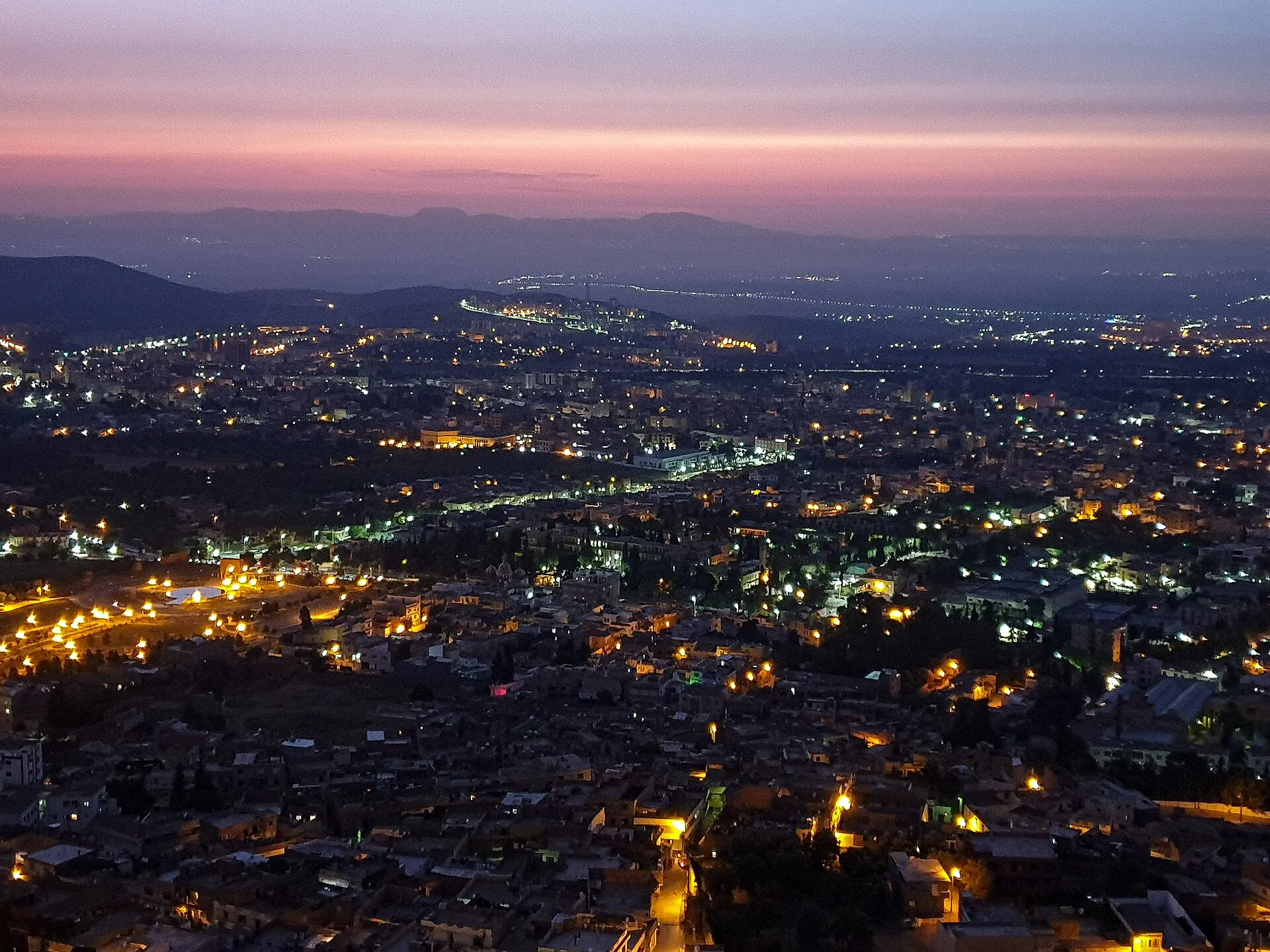 Tlemcen au crépuscule, vue d’en haut : la ville s’allume dans la plaine, sous des montagnes bleutées et un ciel rose